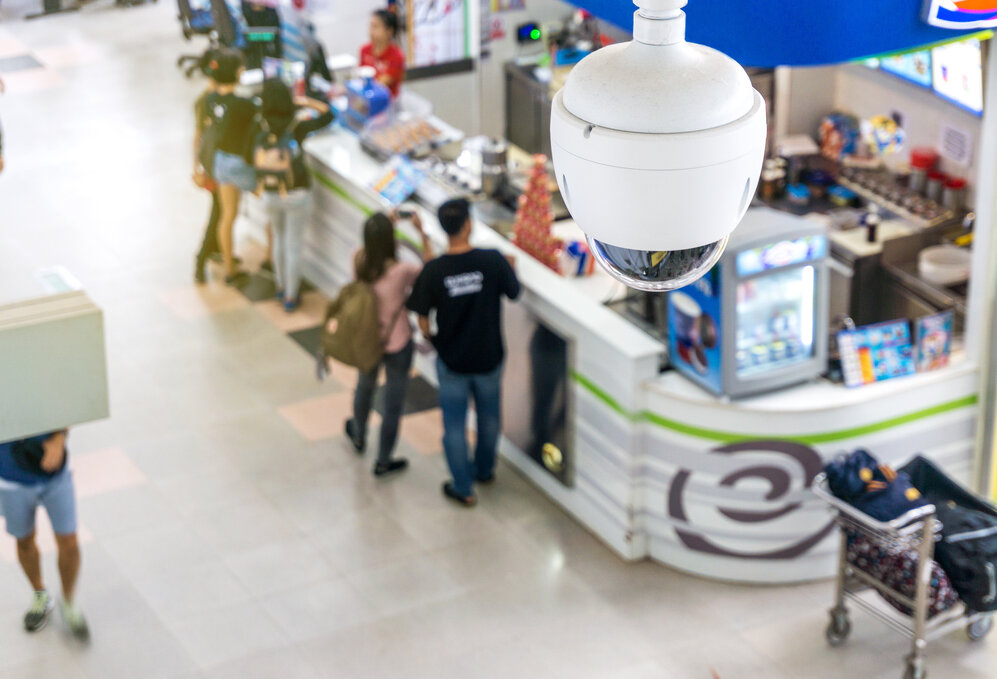 Picture of a white dome security camera above a store sales floor with customers below